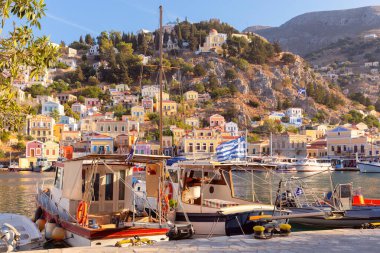 Yunanistan 'ın Symi kentindeki sakin liman sularına demirlemiş renkli balıkçı teknesi, yamaçta geleneksel evleri bulunuyor