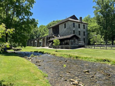 Mitchell, Indiana yakınlarındaki Spring Mill State Park 'ta yeniden inşa edilmiş ve restore edilmiş 1800 Pioneer Village' daki tarihi değirmen ve değirmen mavi gökyüzü ve yeşil çimen alanı olan bir derenin yanında..