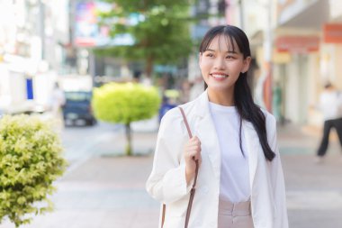 Beyaz gömlek ve omuz çantası giyen Asyalı, kendine güvenen genç iş kadını, işe giderken mutlu bir şekilde gülümsüyor..