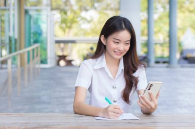 Young Asian woman student in uniform using smartphone and writing something about work.There are many documents on the table, her face with smiling in a working at to search information for a study report at university.