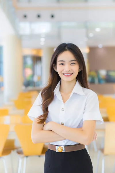 Portrait of an adult Thai student in university student uniform. Asian beautiful girl standing smiling happily and confidently with her arms crossed at the university reading room.