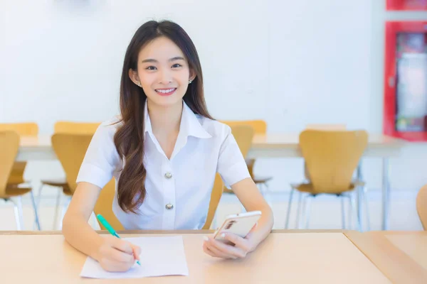 Young Asian woman student in uniform using smartphone and writing something about work.There are many documents on the table, her face with smiling in a working at to search information for a study report at university.