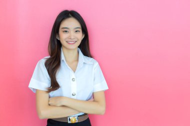 Portrait of adult Thai student. Beautiful Asian young woman student in uniform is smiling and looking at camera with her arms crossed confidently on pink background in the university