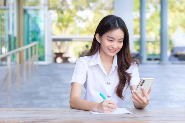 Young Asian woman student in uniform using smartphone and writing something about work.There are many documents on the table, her face with smiling in a working at to search information for a study report at university.