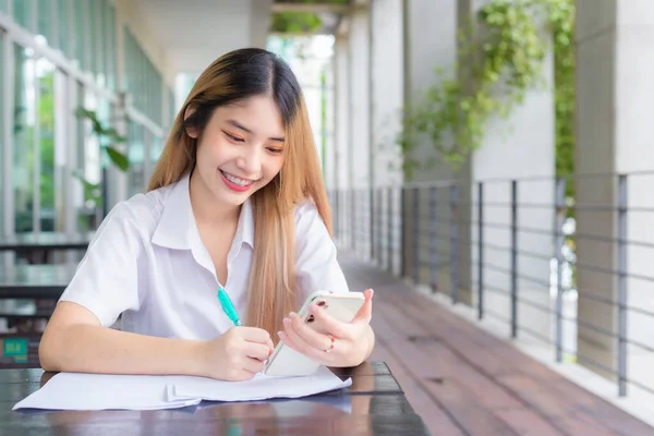 Young Asian woman student in uniform using smartphone and writing something about work.There are many documents on the table, her face with smiling in a working at to search information.