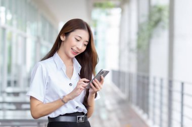 Young Asian beautiful and pretty woman student in uniform using smartphone something about study her face with smiling in a working at to search information at university.
