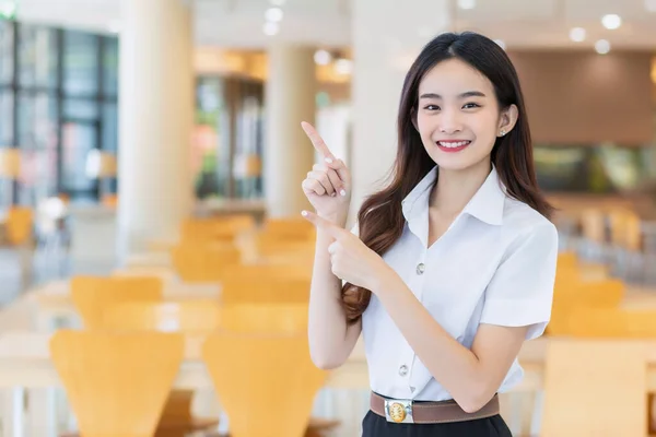 Portrait of an adult Thai student in university student uniform. Asian beautiful girl standing smiling happily and confidently while her hand shows to present something at the university reading room.