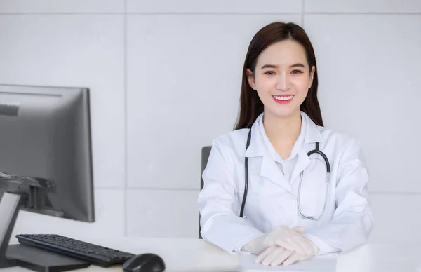 Smiling Asian professional female doctor sitting in examination room at hospital while on table with paper and computer in health concept