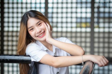 Beautiful young Asian student wearing a Thai university uniform and teething retainer stands smiling cutely in the building during free time while waiting for class.