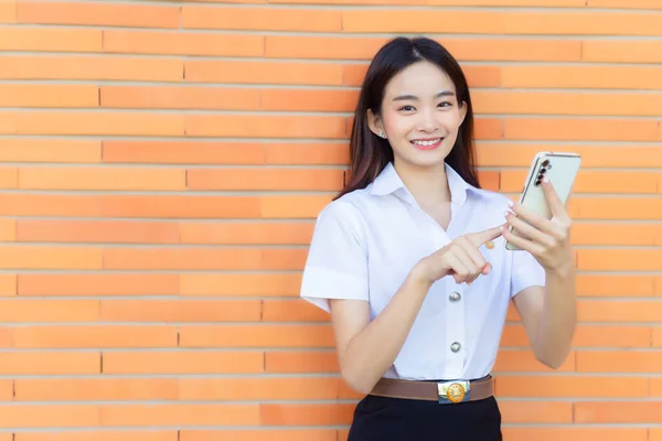 Portrait young Asian beautiful and pretty Thai girl student in uniform while using smartphone she acted excited and looking  camera with brick walls as background in university.