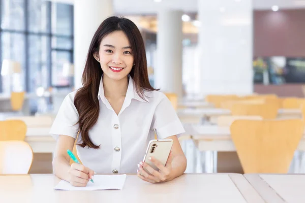 Young Asian woman student in uniform using smartphone and writing something about work. There are many documents on the table, her face with smiling in working at to search information for study at the university reading room.