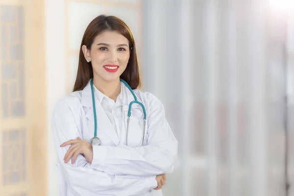 在医院的诊疗室里 专业的年轻亚洲女医生抱着胳膊高高兴兴地站在一起笑着 穿着白色长袍和听诊器 新的正常和保健概念 — 图库照片