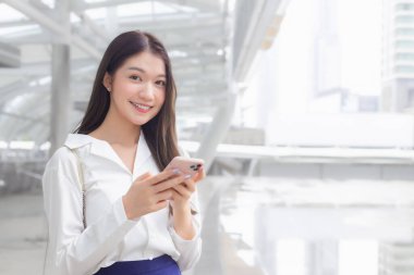 Genç Asyalı kendine güvenen iş kadını ofise ya da iş yerine gidiyor. Kameraya baktığı yer, şehir merkezli iş binalarıyla dolu büyük bir şehirde akıllı telefonu elinde tutuyor..