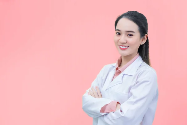 黒い長い髪をしているアジアの女性医師は ヘルスケアのオフィスルームで腕のクロス 桃の色の背景に隔離された新しい通常の概念ながら 均一な白いラボコートを身に着けている — ストック写真