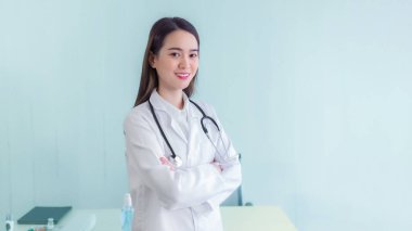 Kollarını kavuşturmuş genç profesyonel Asyalı kadın doktor sağlık hizmetlerinin muayene odasındaki muayene odasında üniforma beyaz bornoz ve steteskop giyiyor. Yeni normal sağlık kontrolü ve koronavirüs koruma kavramı..