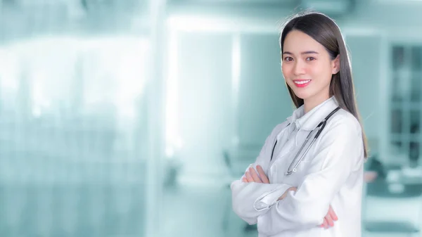 病院の診察室で白いローブと聴診器を着たカメラを見て 腕を組んで立っているプロのかなりアジア系の若い女性医師が笑顔を交わした 健康診断は病気や健康の概念を防ぐ — ストック写真