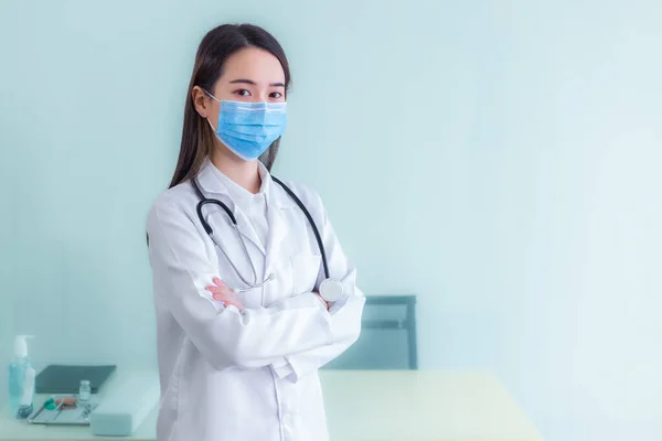 黒髪のプロの若いアジア人女性医師は 彼女が病院の健康診断室で腕のクロスながら 彼女は医療面のマスクを身に着けていると一様として聴診器や白衣を身に着けています — ストック写真