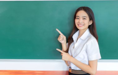 Portrait of adult Thai student. Beautiful Asian young woman student in uniform is smiling and looking at camera to present something confidently with blackboard, green as background in classroom at the university. clipart