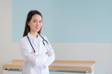 Asyalı profesyonel bayan doktor beyaz bornoz ve steteskop giyip kollarını kavuşturup mutlu mesut gülümseyerek hastanedeki çocuk ya da genç çocuk muayene odasında duruyor.. 