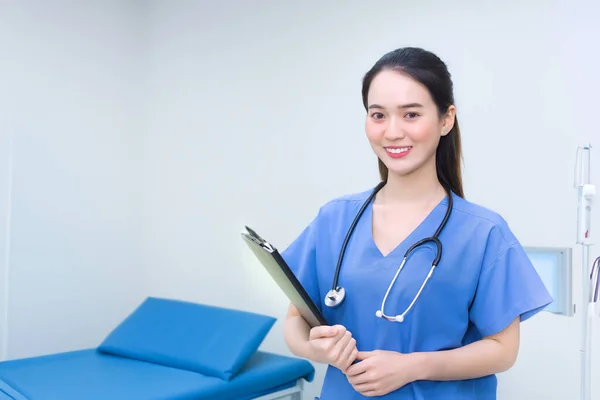 专业的年轻亚洲女医生站在医院的病房里 穿着军服蓝色的实验室衬衫 手里拿着病人的文件 微笑着 保健概念 — 图库照片