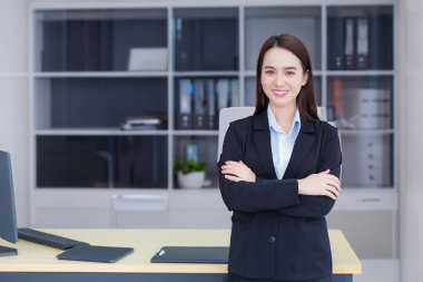 Uzun saçlı, Asyalı, profesyonel bir iş kadını. Mavi gömlekli, siyah bir takım elbise giyiyor. Ofis odasında kollarını kavuşturup gülümsüyor..