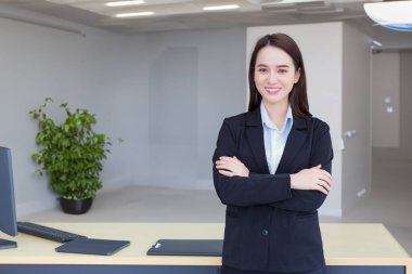 Uzun saçlı Asyalı profesyonel bir kadın ofis odasında kol kola girerken ve kendine güvenen gülümserken mavi gömlekli siyah takım elbise giyiyor..