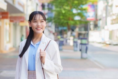 Beyaz gömlek ve omuz çantası giyen kendine güvenen genç Asyalı kadın işe giderken mutlu bir şekilde gülümsüyor..