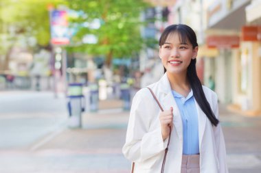 Beyaz gömlek ve omuz çantası giyen kendine güvenen genç Asyalı kadın işe giderken mutlu bir şekilde gülümsüyor..