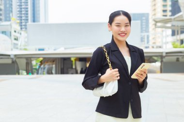 Koyu mavi takım elbiseli genç Asyalı profesyonel kadın arka planda şehir binalarıyla işe giderken akıllı telefonunu elinde tutuyor..