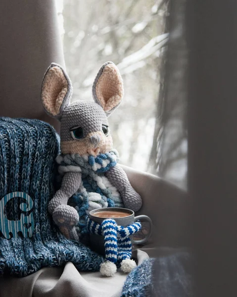 Handmade knitted toy bunny rabbit hare sitting near the window with big cup of hot chocolate, winter holidays concept