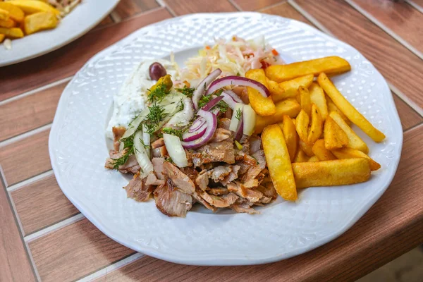 Gyros, Greek dish from sliced meat roasted on a turning spit, served with French fries, coleslaw, tzatziki and onions on a white plate, wooden outdoor table, selected focus, narrow depth of field