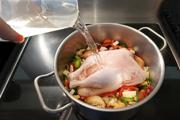 Whole raw chicken in a pot with chopped vegetables is poured with cold water for cooking a healthy soup against cold and flu, selected focus, narrow depth of field