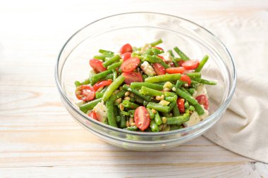 Green bean salad with tomatoes, pine nuts and feta cheese in a glass bowl on a light wooden table, Mediterranean snack or antipasto, copy space, selected focus, narrow depth of field