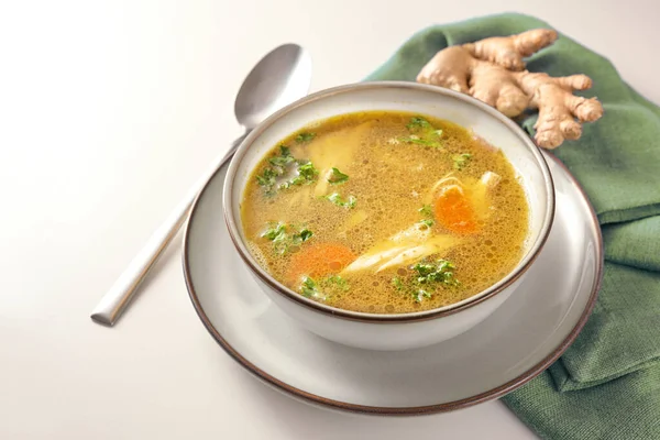 Homemade chicken soup with vegetables, ginger and parsley in a bowl, green napkin and spoon on a gay white table, copy space, selected focus, narrow depth of field