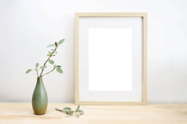 Empty wooden picture frame with passe-partout as mock-up and a vase with sage twig on a light table or desk in front of a white wall, minimalist interior decoration, copy space, selected focus