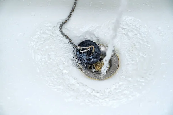 Water running in a white wash basin or bathtub with an old black rubber plug and a rusty drain, copy space, selected focus, narrow depth of field