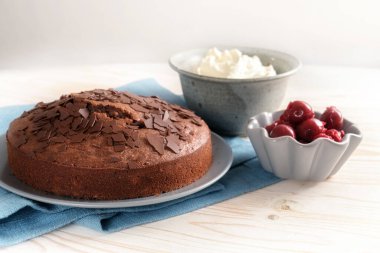 Chocolate cake and bowls with cherries and whipped cream on a blue napkin and a light wooden table, copy space, selected soft focus, narrow depth of field
