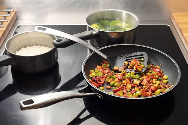 Diced vegetables are stir-fried with spices in a frying pan, pots with rice and broccoli behind them on the black stovetop, Asian style cooking, copy space, selected focus, narrow depth of field