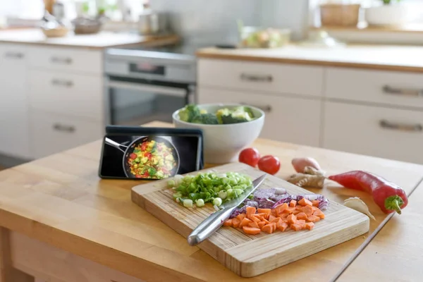 Cooking by using an online app. Smartphone, diced vegetables and a knife on a wooden board, blurred kitchen in the background, selected focus, narrow depth of field