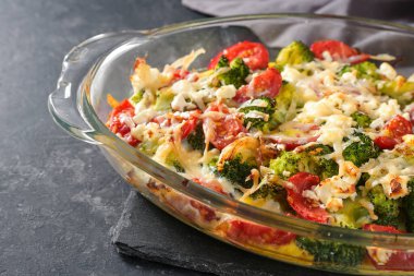 Vegetable dish from broccoli, tomatoes, onions, feta and cheese in a glass casserole on a gray slate table, copy space, selected focus, narrow depth of field