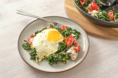 Fried egg on spinach with tomatoes and cheese served on a plate on a light painted wooden table, homemade healthy dish, copy space, selected focus, narrow depth of field