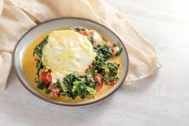 Spinach dish with tomatoes, ham, cheese and a baked egg in a spicy sauce on a plate and a light wooden table, copy space, selected focus, narrow depth of field