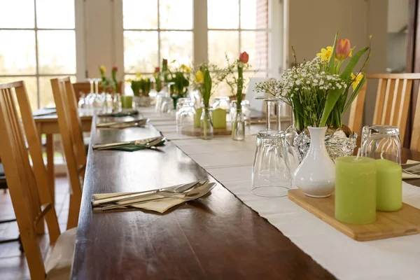 春の花とディナーパーティーのために飾られた大きな木製のテーブル 緑のキャンドル ダイニングルームでのライトテーブルランナー上の様々な飲料ガラス 選択されたフォーカス フィールドの非常に狭い深さ — ストック写真