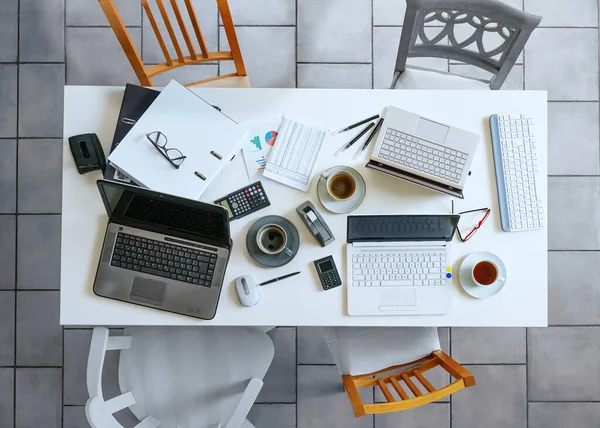 Top view of a small white meeting table with laptops, coffee, calculator, papers, ring binder and four casual chairs on a gray tiled floor business, high angle view form above, selected focus