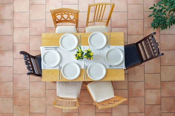 Top view of a place setting with six white plates, cutlery and yellow daffodil flowers on a light wooden table and six different chairs on a terracotta tiled floor, selected focus