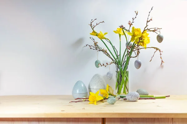 春天的装饰 用一束水仙花和玻璃瓶中的枝条装饰 用木制餐具柜上的人造蓝色灰色彩蛋靠着轻墙 复制空间 选定焦点 — 图库照片