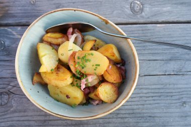 Kızarmış patates, soğan, domuz pastırması ve soğan süslemesi mavi gri ahşap bir masada seramik bir kasede, yukarıdan yüksek açılı görüş, kopyalama alanı, seçilmiş odak alanı, dar alan derinliği