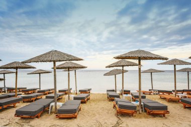Yumuşak yastıklı ahşap plaj şezlongları ve saman güneş şemsiyeleri Yunanistan 'da bir turizm beldesinde bulutlu mavi gökyüzüne, kopya alanına ve seçilmiş odak noktasına karşı.