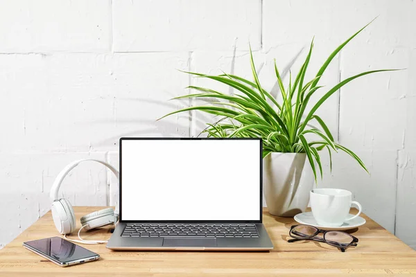 Wooden desk with open laptop mockup, headphones, phone, glasses and plant in front of a white painted wall, office, business work and network concept, copy space, selected focus