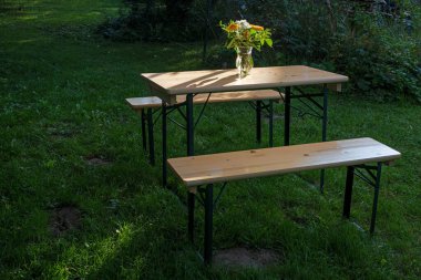 Çiçek buketi olan basit ahşap masa ve güneşli bir günde doğal bir bahçedeki çayırlarda banklar, cennet gibi bir yaz sahnesi, kopya alanı, seçilmiş yumuşak odak, dar bir alan derinliği.